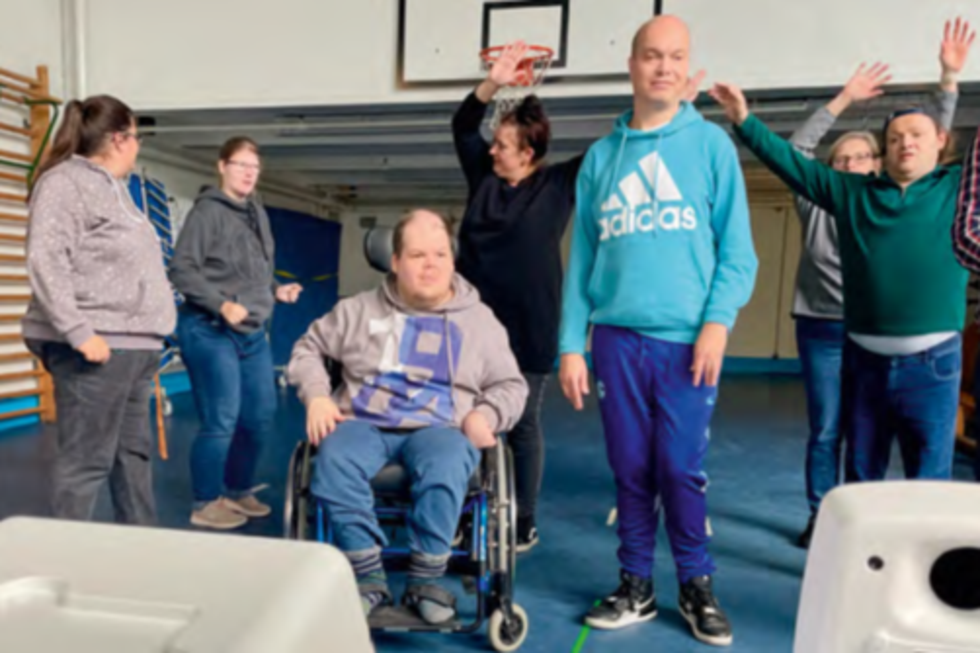 Mehrere Personen befinden sich in einer Turnhalle, eine Person sitzt im Rollstuhl, andere stehen oder heben die Arme.