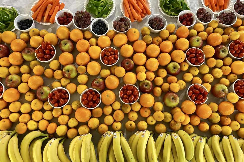 Verschiedene Obst- und Gemüsesorten, darunter Bananen, Äpfel, Orangen, Zitronen, Tomaten, Karotten, Paprika, Trauben und Beeren, sind in Reihen auf einem Tisch angeordnet.