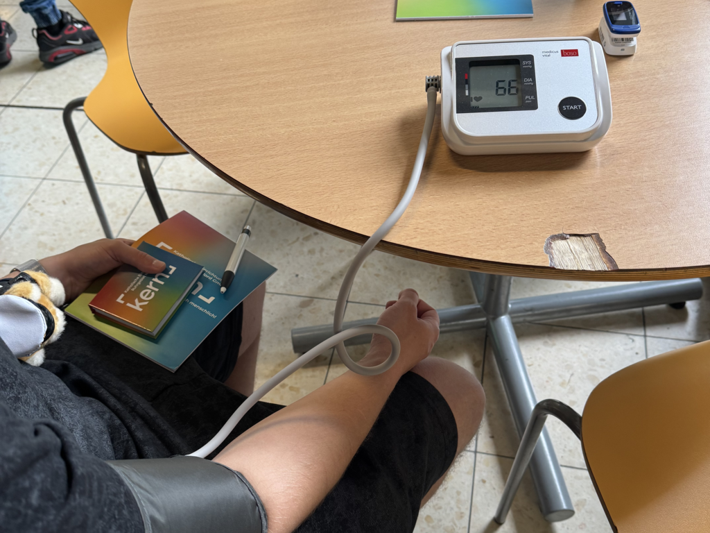 Eine Person misst den Blutdruck mit einem elektronischen Messgerät am Arm, während sie an einem Tisch sitzt.