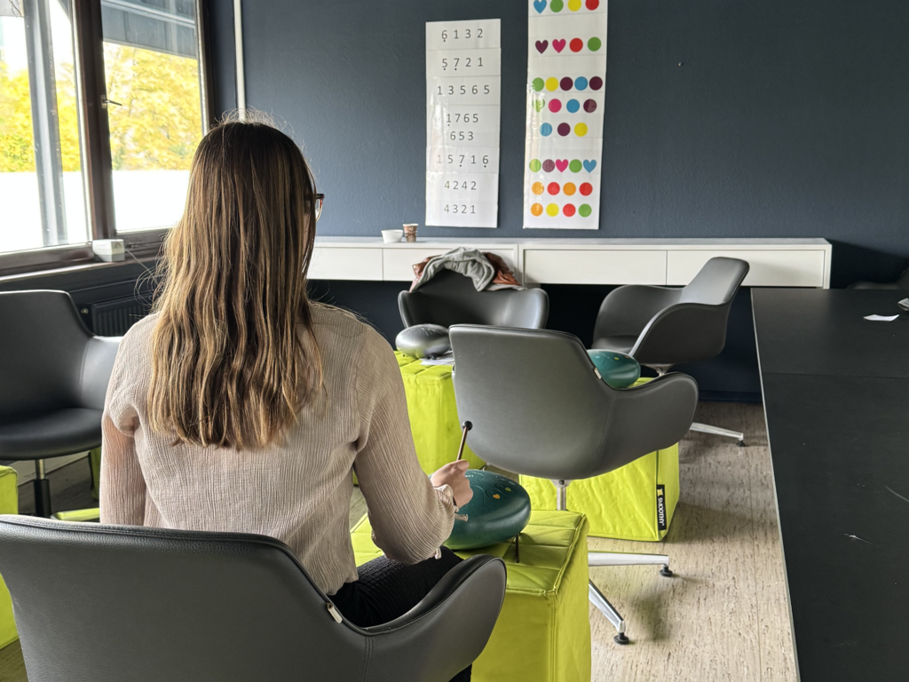 Eine Person sitzt auf einem grauen Stuhl und hält einen Schlägel über ein grünes Musikinstrument, im Hintergrund sind weitere Stühle, gelbe Sitzwürfel und zwei Poster mit Zahlen und bunten Punkten an der Wand.