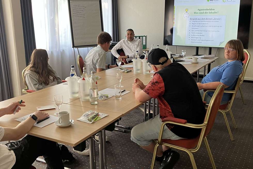 Sechs Personen sitzen an einem Konferenztisch in einem Besprechungsraum, auf dem Tisch stehen Getränke und Gläser, im Hintergrund sind ein Flipchart und ein großer Bildschirm mit einer Präsentation.