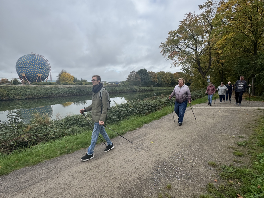 Mehrere Personen gehen mit Nordic-Walking-Stöcken auf einem Weg an einem Fluss entlang, im Hintergrund steht ein kugelförmiges Gebäude.