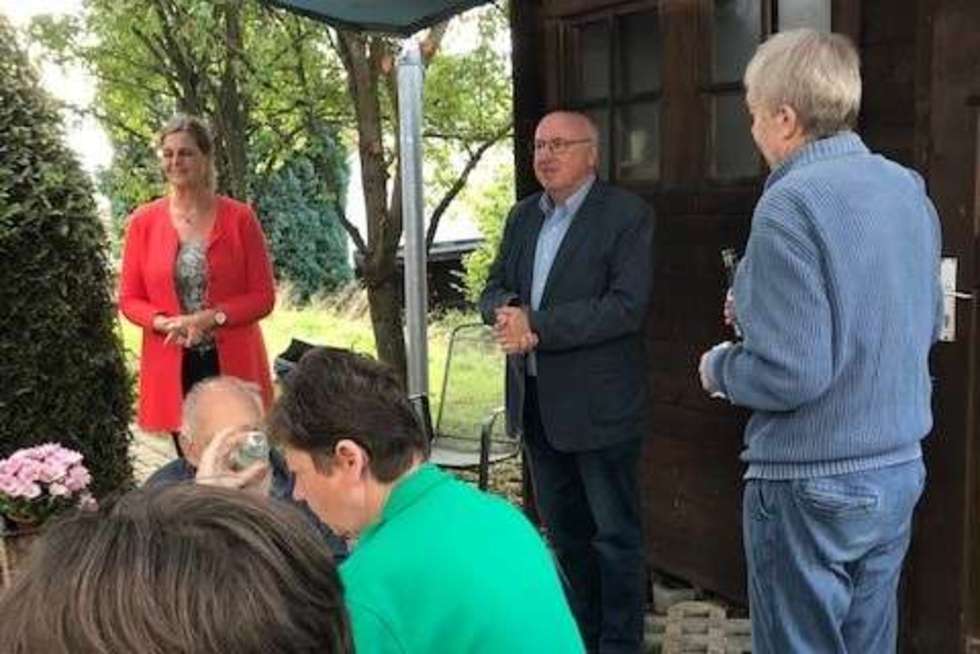 Mehrere Personen befinden sich im Freien vor einem Holzhaus, einige sitzen an einem Tisch, andere stehen und sprechen miteinander.