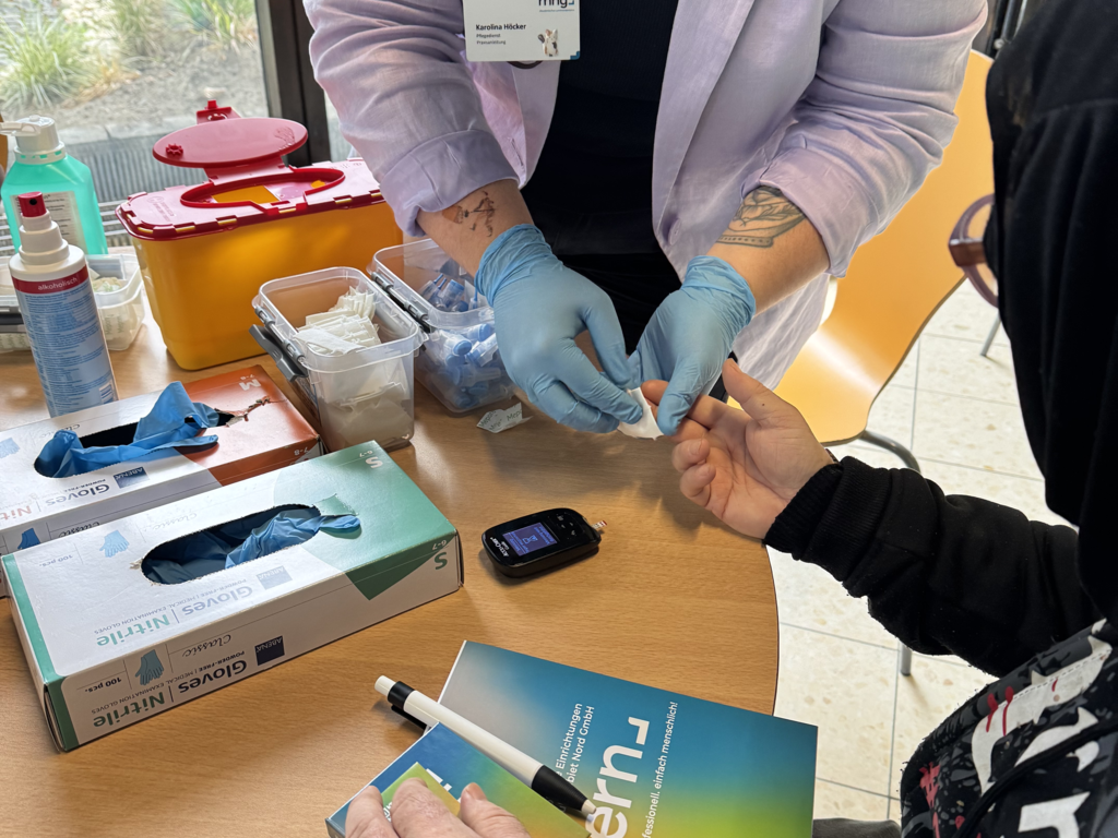 Eine Person mit Handschuhen nimmt einer anderen Person Blut am Finger ab, auf dem Tisch liegen medizinische Utensilien und ein Blutzuckermessgerät.