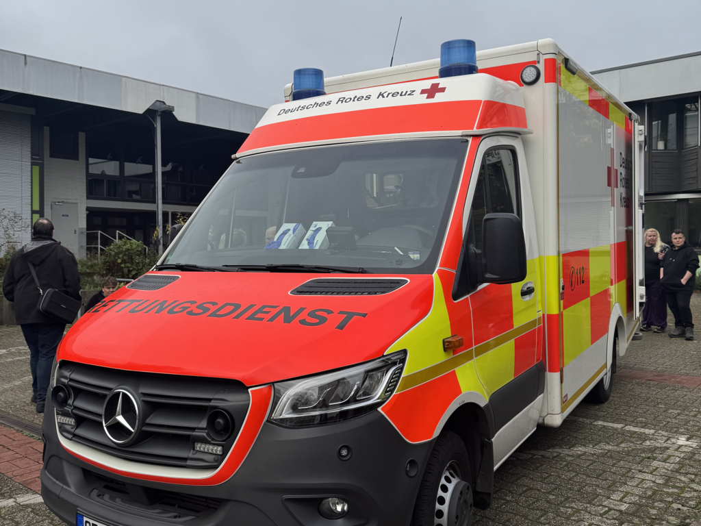 Ein Rettungswagen mit der Aufschrift „Deutsches Rotes Kreuz“ steht auf einem Parkplatz, im Hintergrund sind mehrere Personen und Gebäude.