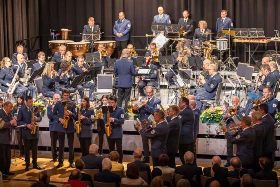 Mehrere Personen in Uniformen spielen auf einer Bühne verschiedene Blasinstrumente vor einem sitzenden Publikum.