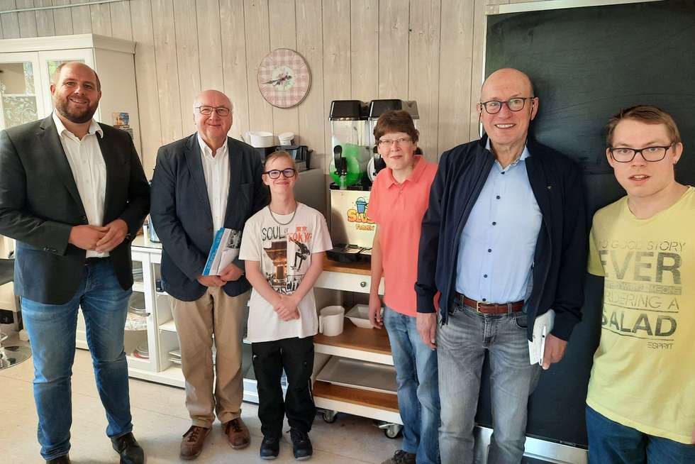 Sechs Personen stehen nebeneinander in einem Raum mit Küchenmöbeln und einer Tafel.